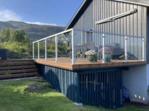 Glassrekkverk med hvite aluminiumstolper rundt treterrasse med utsikt over frodig fjellandskap.