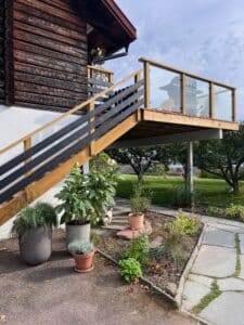 Høyt plassert altan med rekkverk i tre og klart glass ved laftet husfasade – utsikt over hage og grøntområde.