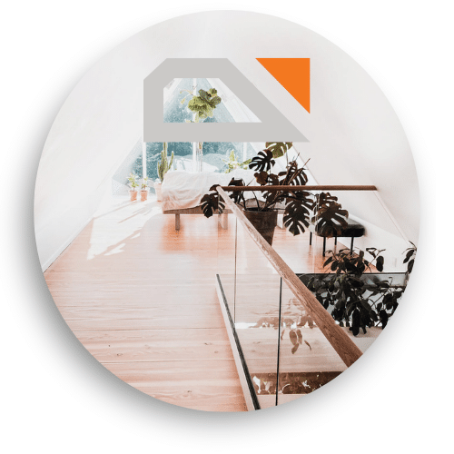 Innendørs bilde som viser et glassrekkverk med tredetaljer ved en hems. Rekkverket strekker seg langs tregulvet, og dagslyset fra vinduet gir et lyst og åpent inntrykk. Flere grønne planter skaper en harmonisk og frodig stemning.