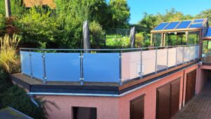 Terrasse med frostet glassrekkverk og runde stolper i rustfritt stål, omgitt av hage og vinterhage.