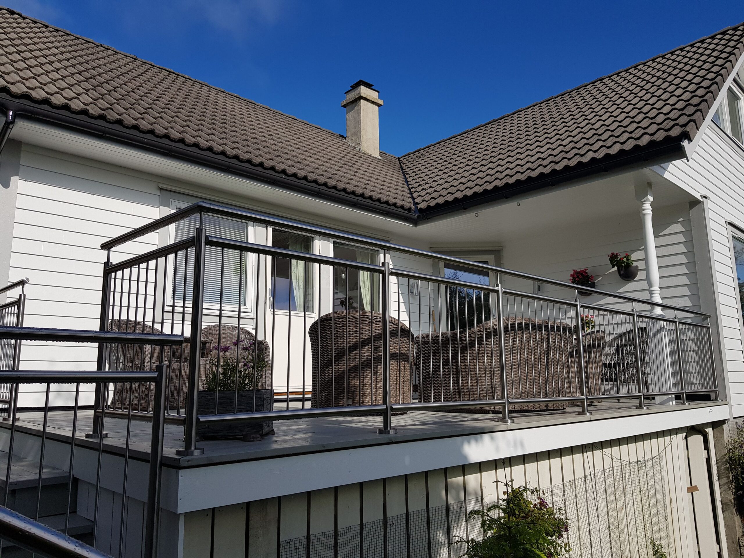Vinklet rekkverk i rustfritt stål rundt hevet veranda med grå terrassebord, sittegruppe og blomsterkrukker foran hvit fasade