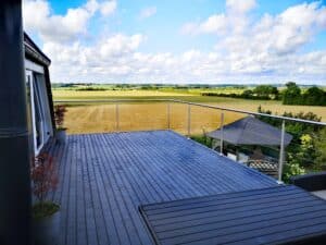 Rekkverk i rustfritt stål med vaierløsning på altan med mørkgrå terrasse – utsikt over jorder og grønne marker.