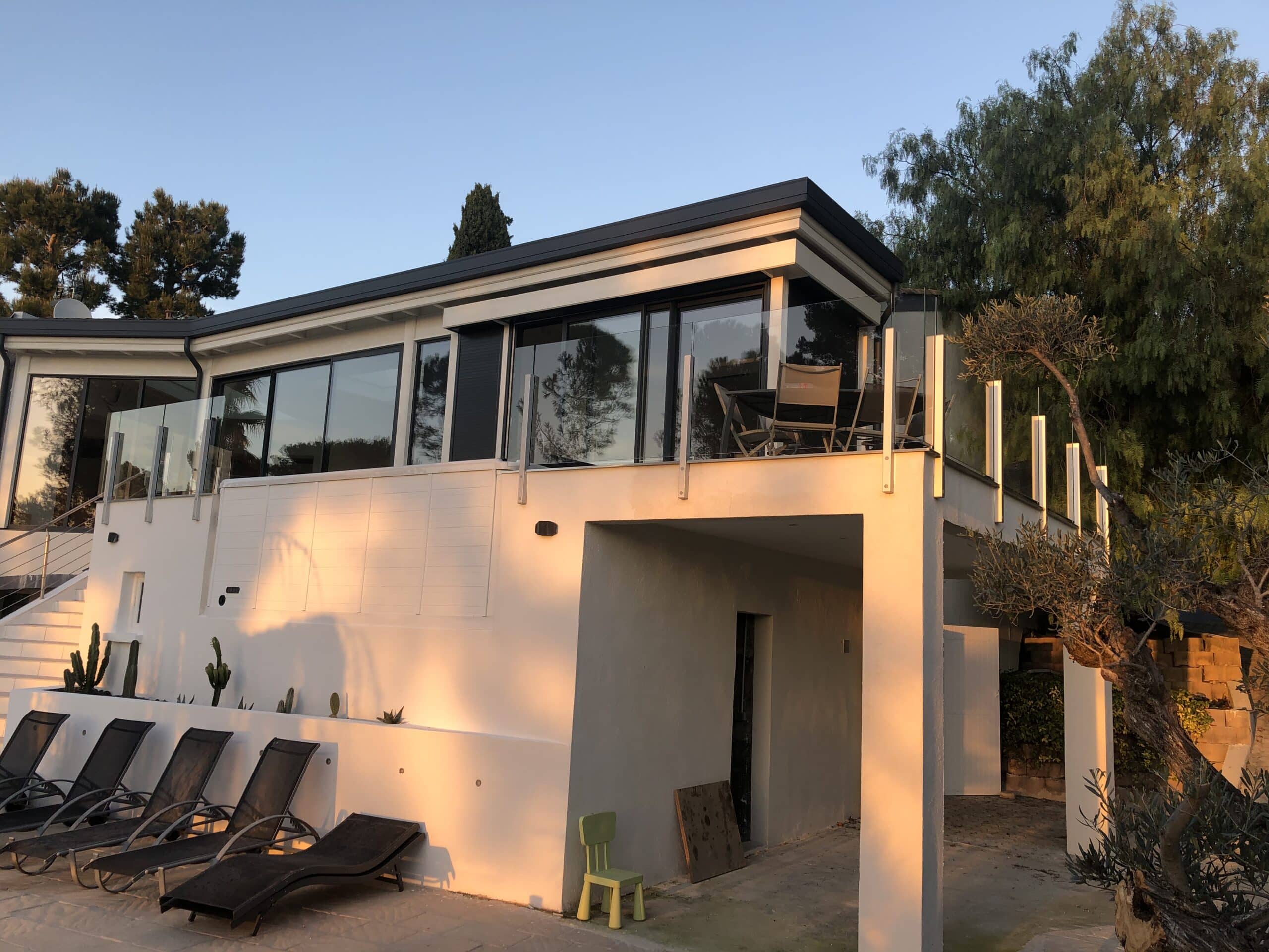 Terrasse med klart glass og aluminiumsrekkverk, badet i gyllen solnedgang med grønne omgivelser og loungestoler.