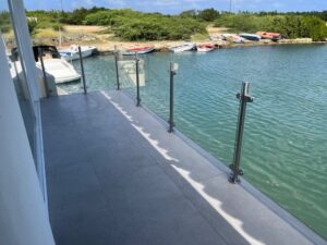 Glassrekkverk med rustfrie stolper langs brygge med utsikt til småbåter og grønt landskap i bakgrunnen.