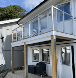 Glassrekkverk med hvite aluminiumstolper montert på balkong med tretak og fasade i naturtre.