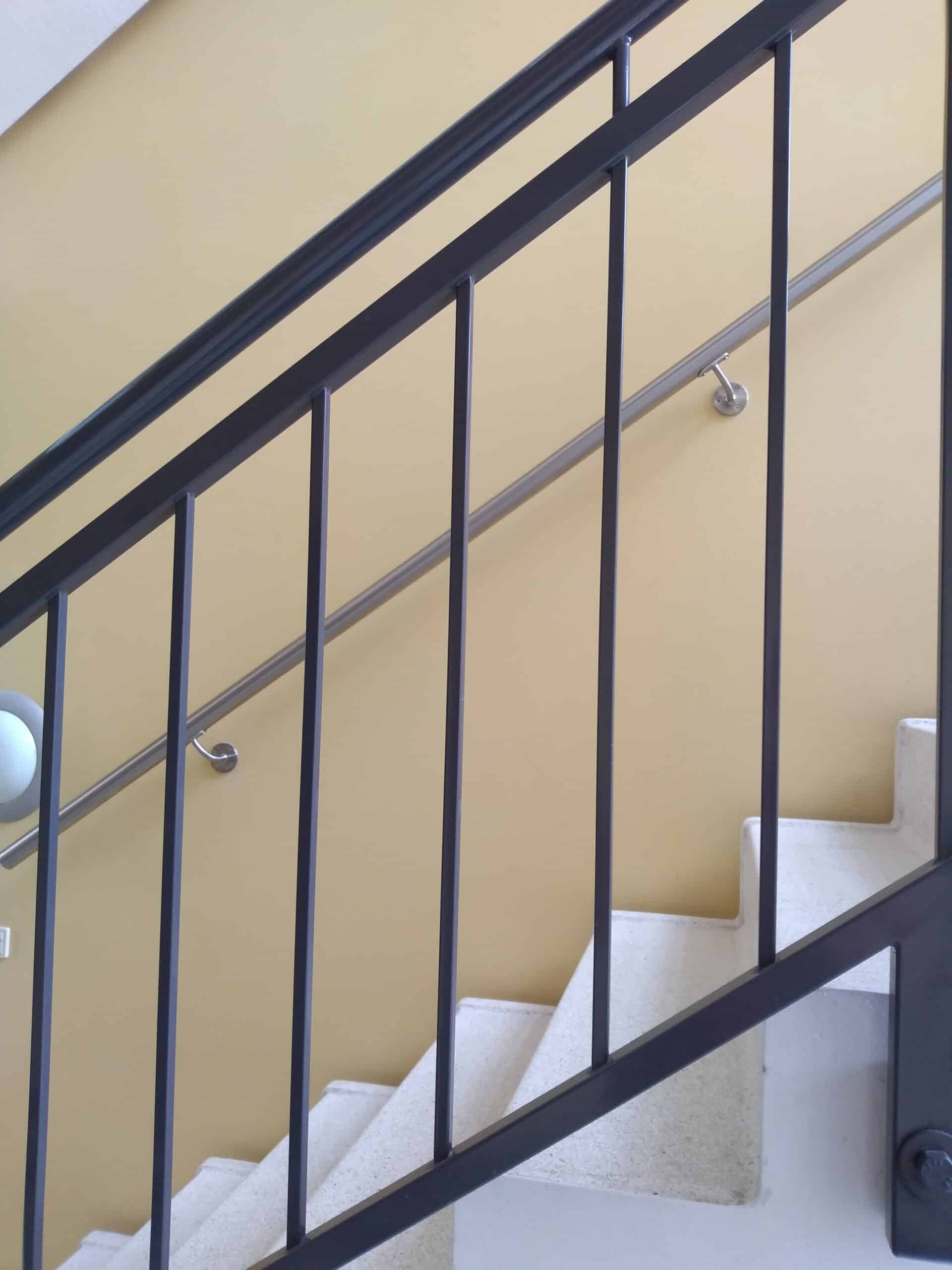 Trapp med svart rekkverk og separat veggmontert håndløper i rustfritt stål for økt sikkerhet.