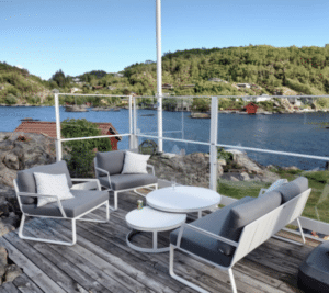 Terrasse med glassrekkverk i hvitlakkert aluminium, plassert på svaberg ved sjøen. Rekkverket har klemfester og transparente glassflater. Hagemøbler i hvitt og grått står på tredekke med utsikt over fjord, røde sjøboder og grønne fjell.