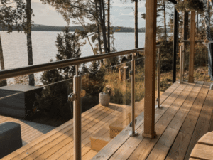 Glassrekkverk med rustfrie stolper og overligger i tre, montert på terrasse i treverk. Glasspanelene holdes på plass med klemfester. I bakgrunnen vises innsjø omgitt av skog og en trapp i tre som fører ned til vannet.